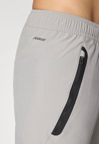 Bermudas deportivas de color gris claro con cintura elástica, bolsillo lateral con cremallera delineado en negro y el texto "AEROREADY" cerca de la cintura.