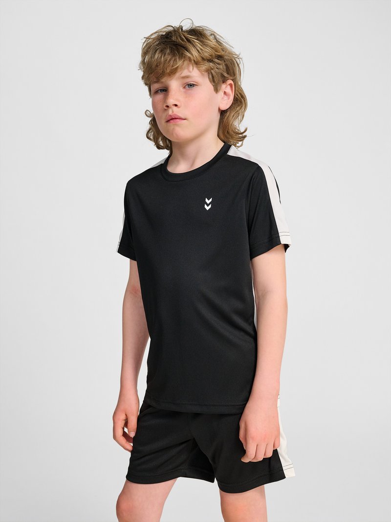 Blonder Junge steht, trägt schwarze Sportkleidung mit weißen Streifen an Ärmeln und Shorts, neutraler Gesichtsausdruck, grauer Hintergrund.