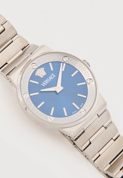 Ασημένιο μεταλλικό ρολόι χειρός με μπλε καντράν, ασημένιους δείκτες ώρας, λογότυπο Versace και έμβλημα Μέδουσας στη θέση 12, δείχνοντας ώρα 11:59.