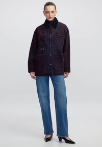 Vrouw in een donkere paarse corduroy jas met grote zakken over een kastanjebruine coltrui, blauwe rechte jeans en zwarte hakken.