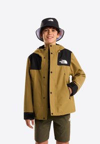 Khaki regnjakke med sorte accenter, udstyret med hætte, trykknapper og et logo mærke. Kombineret med grønne shorts. Sort bøllehat.