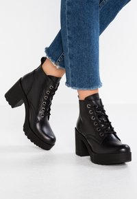 Black lace-up ankle boots made of smooth leather, featuring a chunky high heel and a ridged sole. Jeans with frayed hems complement the boots.