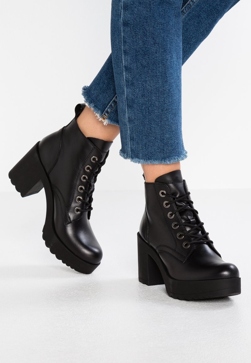Black lace-up ankle boots made of smooth leather, featuring a chunky high heel and a ridged sole. Jeans with frayed hems complement the boots.