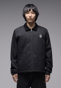 Μαύρο μπουφάν με φερμουάρ και γιακά, με κυκλικό λογότυπο στο αριστερό στήθος και μακριά μανίκια με μανσέτες. Λείος υφασμάτινος υφή.