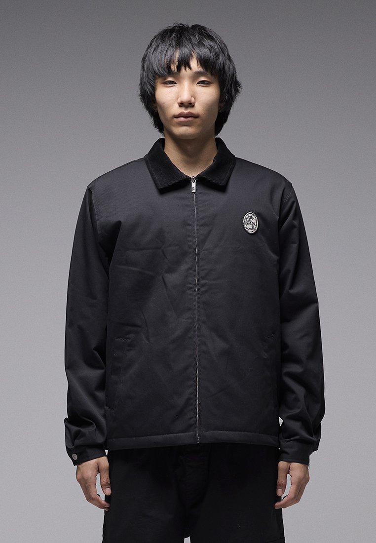 Μαύρο μπουφάν με φερμουάρ και γιακά, με κυκλικό λογότυπο στο αριστερό στήθος και μακριά μανίκια με μανσέτες. Λείος υφασμάτινος υφή.