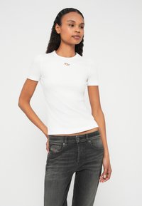 Λευκό ριμπ κοντομάνικο τοπ με κεντρική λεπτομέρεια λογότυπου, συνδυασμένο με σκούρα γκρι τζιν τζιν που εμφανίζει ελαφρώς ξεθωριασμένο εφέ και διακοσμητικές ραφές στις τσέπες.