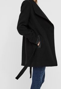 Schwarze Jacke mit breitem Kragen, langen Ärmeln und Seitentaschen. Verfügt über einen Gürtel in der Taille und strukturierte Lederelemente an den Bündchen.