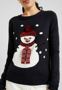 Maglione blu navy con una grafica di un pupazzo di neve bianco, dotato di un cappello rosso, sciarpa e pon-pon, accentuato da fiocchi di neve. Morbida texture a maglia.