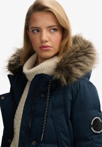 Giacca puffer blu navy con cappuccio foderato in pelliccia, indossata sopra un maglione crema e con una patch sulla manica. Tessuto strutturato e zip visibili.