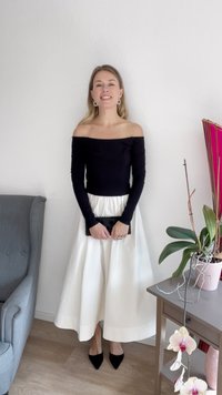 Svart off-shoulder topp, vit hel kjol, svart kuvertväska och spetsiga svarta klackar. Inomhusmiljö med en växt och dekorativa detaljer.