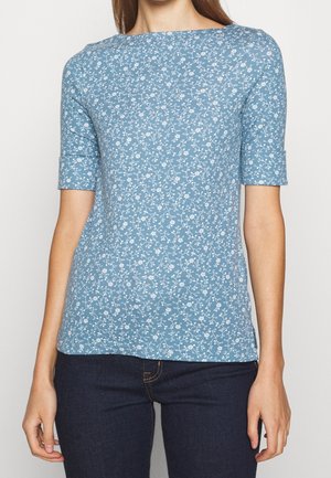 Kobieta ubrana w niebieską bluzkę z krótkim rękawem w biały wzór kwiatowy oraz ciemnoniebieskie, dopasowane dżinsy, stojąca na prostym białym tle.