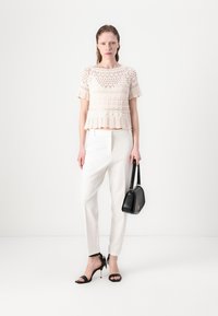 Crochet top v svetlobeige farbe s krátkymi rukávmi a peplum golierom, v kombinácii s upravenými bielymi nohavicami a čiernymi slingback lodičkami, drží čiernu tašku.