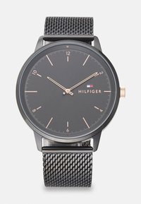 Black analog watch with a round face, minimalist design, rose gold hour markers, and mesh strap. Brand name "HILFIGER" displayed.