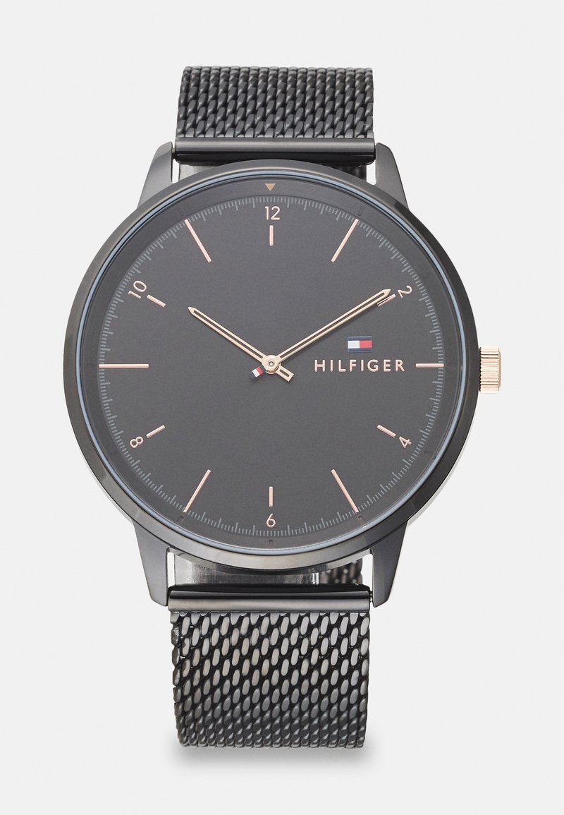 Black analog watch with a round face, minimalist design, rose gold hour markers, and mesh strap. Brand name "HILFIGER" displayed.