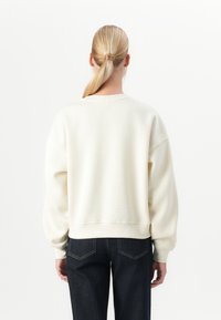 Mujer con el cabello rubio recogido en una coleta baja, vestida con un sudadera holgada color blanco roto y jeans azules oscuros, de pie contra un fondo blanco liso.