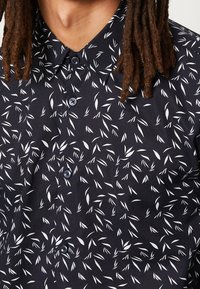 Lyhythihainen nappikauluspaita tummansinisestä kankaasta, jossa on valkoinen lehtikuosi. Klassinen kaulus sinisillä nappeilla. Kevyt rakenne.