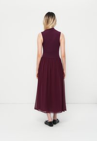 Woman with long hair wearing a sleeveless, high-neck plum dress with a sheer skirt, standing facing a white wall and black flats.
