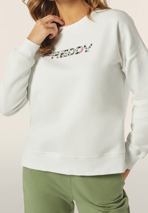 Femme portant un sweat-shirt blanc avec un logo floral "FREDDY" et un pantalon vert clair, main touchant les cheveux près de l'épaule.