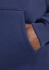 Felpa blu navy con tasca frontale, polsini a costine e una texture morbida. Il dettaglio mostra una mano appoggiata nella tasca.