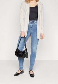 Lichtbeige blazer over een zwarte top, gecombineerd met blauwe skinny jeans en zwarte flats. Een kleine zwarte handtas met hardwaredetails maakt de look compleet.