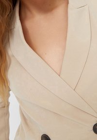 Gros plan d'une femme portant un blazer croisé beige à boutons noirs, avec des cheveux blonds ondulés tombant sur son épaule.