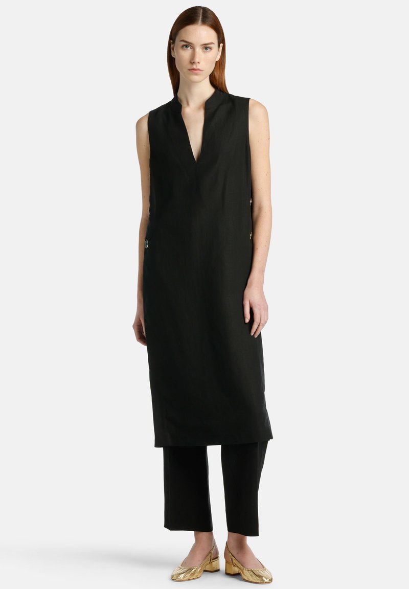 Black sleeveless dress with a deep V-neck, side buttons, paired with black cropped pants and gold heeled shoes, smooth fabric texture.