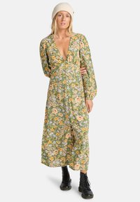 Vestido maxi floral en verde con patrones naranjas y morados, que presenta un escote en V profundo, mangas abullonadas y detalles de botones. Combinado con botas negras.