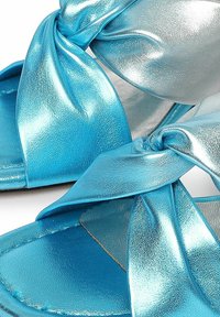Sandali slide metallici blu con un design a strap annodati, texture liscia e soletta imbottita. Gli accenti includono una finitura argento a contrasto.