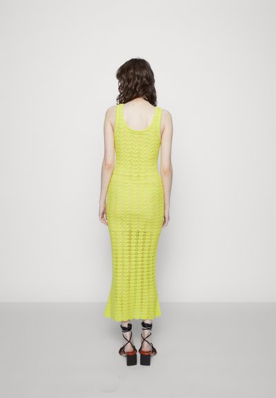 Vestido maxi de malha leve na cor amarela brilhante. Apresenta um design sem mangas, silhueta ajustada e bainha com bordas recortadas. Usado com saltos pretos com tiras.