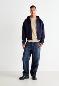 Sweatshirt à capuche navy avec fermeture éclair, t-shirt beige en dessous, associé à un jean bleu ample et des chaussures noires. Deux poches avant sur le sweatshirt.