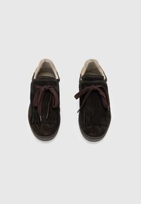 Un paio di mocassini in suede marrone scuro con dettagli di frange e lacci legati, esposti su uno sfondo grigio chiaro.