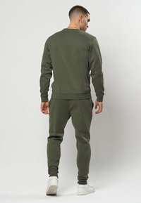 Bărbat în picioare, cu fața spre spate, purtând un hanorac verde măsliniu, pantaloni de trening asortați și adidași albi, pe un fundal simplu, deschis la culoare.