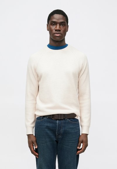 Lichtbeige geribbelde trui met ronde hals, gedragen over een blauwe coltrui, gecombineerd met donkerblauwe spijkerbroek en een bruine riem.