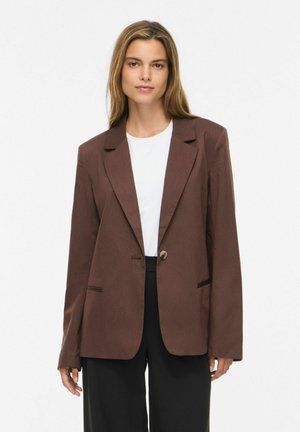 Kvinde med glat lysebrunt hår iført brun enkeltknappet blazer, hvid skjorte og sorte bukser mod en ensfarvet lys baggrund.