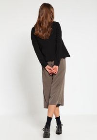 Nainen seisoo kädet ristissä selän takana, päällä musta takki, taupe-harmaat culottes-housut, mustat sukat ja mustat kengät valkoista taustaa vasten.