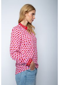 Langärmliges Hemd mit geometrischem Muster in Pink, Rot und Weiß. Rotes Taillenbündchen und Kragen mit der Aufschrift "LOVE". Der Stoff wirkt leicht und hat eine glatte Textur.