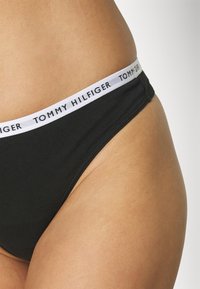 Musta puuvillabikini valge vööga, millel on musta värviga trükitud "TOMMY HILFIGER". Kangas on siledat tekstuuri ja sobib hästi.