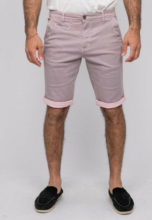 Homme portant un short rose clair retroussé, des chaussures noires sans lacets et une chemise blanche, debout devant un fond uni.