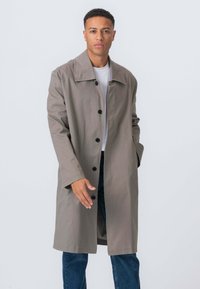 Grå bomulds trenchcoat med bred krave, lange ærmer og seks sorte knapper; båret over en hvid skjorte og blå jeans.