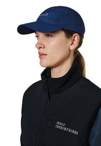 Vrouw in profiel met een donkerblauwe pet met ventilatiegaatjes en een zwarte jas met rits met "HALO" en nummers gedrukt op de borst.