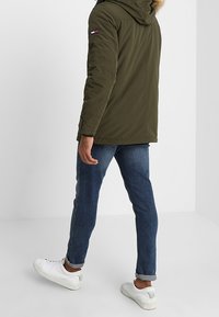 Olivgrön parkajacka med foder av päls i huvan, dragkedjestängning, sidofickor och logotypmärke på ärmen. Bärs med blå jeans och vita sneakers.