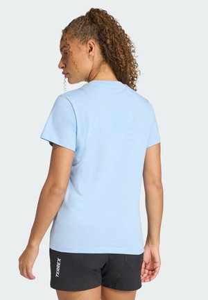 Vrouw met krullend haar die een lichtblauw shirt met korte mouwen en zwarte Terrex-shorts draagt, van achteren gezien tegen een effen achtergrond.