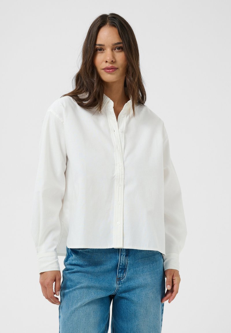 Femme portant une chemise blanche ample à boutons avec manches longues et un jean en denim bleu sur un fond clair uni.