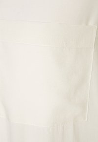 Tissu couleur crème à texture douce, avec une grande poche rectangulaire à l'avant et des détails de couture subtils.