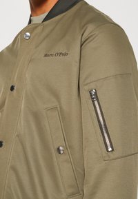 Oliivinvihreä bomber-takki, jossa vetoketjullinen hihasuoja, painonappisuljin ja kirjailtu logo. Takissa on teksturoitu, sileä kangas.