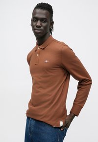 Bruine polo met lange mouwen, een kraag, een knoopsluiting met twee knopen en een geborduurd logo. Gemaakt van gestructureerde stof, met zijopeningen.
