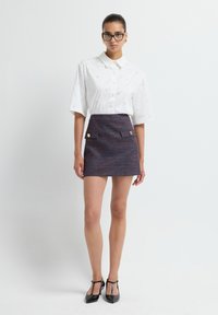 Perempuan yang mengenakan kacamata, blus lengan pendek putih dengan mutiara kecil, rok mini tekstur gelap dengan kancing emas, dan sepatu hak T-straps hitam.