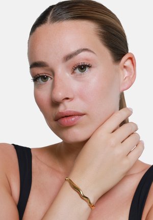 Bracelet large en or ondulé au poignet, présentant un métal lisse et réfléchissant. Le teint est neutre contre des vêtements noirs et un fond minimaliste.