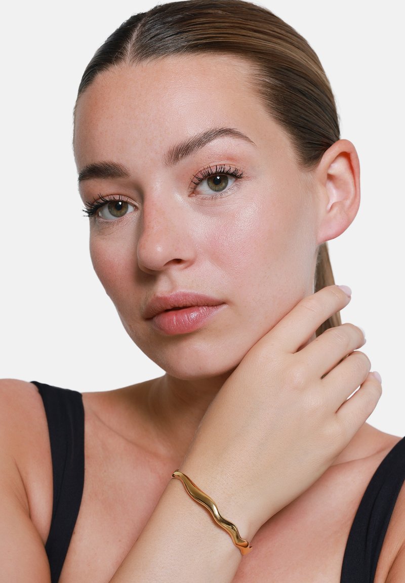 Gouden golvende manchetarmband om de pols, met glad, reflecterend metaal. Huidskleur is neutraal tegen zwarte kleding en een minimalistische achtergrond.