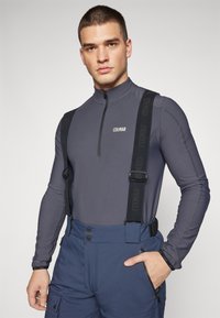 Gray zip-up base layer with high collar, paired with navy blue ski pants featuring adjustable black straps, buttons, and side pockets.